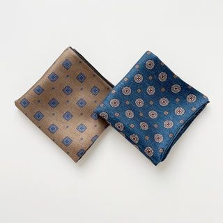 Two luxury silk neckerchiefs displayed side by side - denim blue with circular medallion pattern and taupe with diamond motif, both hand-rolled.