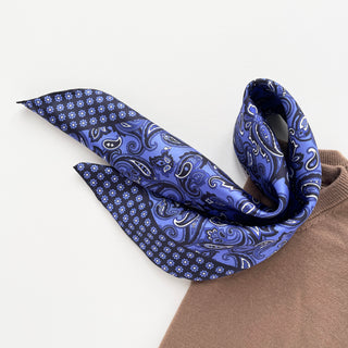 Navy blue silk neckerchief with paisley pattern and floral border, elegantly draped over light brown fabric showing luxurious sheen and hand-rolled edges