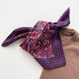 Plum red silk neckerchief with navy blue border featuring classic paisley pattern and floral trim, styled with beige sweater