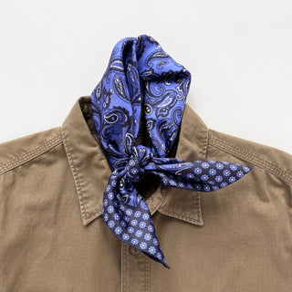 Navy blue silk neckerchief with paisley pattern and floral edge detail, tied stylishly around the collar of a tan button-up shirt