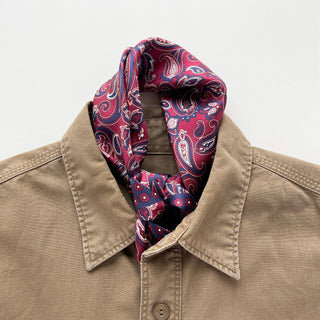 Plum red silk neckerchief with navy blue paisley pattern elegantly tied around the collar of a tan button-up shirt, showcasing luxurious texture and styling