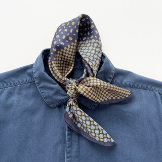 Navy silk neckerchief with paisley and geometric patterns in gold and blue tones, elegantly tied around a blue shirt collar