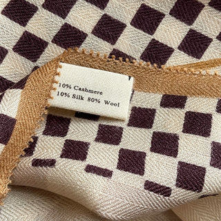 Close-up of scarf fabric showing cream and dark brown checkerboard pattern with golden trim and material label indicating 10% cashmere, 10% silk, 80% wool blend.