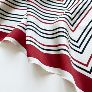 Luxurious silk scarf with geometric pattern featuring red and navy stripes on off-white background, showing fabric's smooth texture and elegant drape.