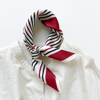 Square silk bandana scarf with red, navy and white striped pattern, tied elegantly around collar of white blouse, showing luxurious sheen