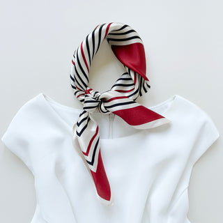 Silk bandana scarf with geometric pattern in red, navy and white stripes, elegantly tied around white garment neckline against light background.
