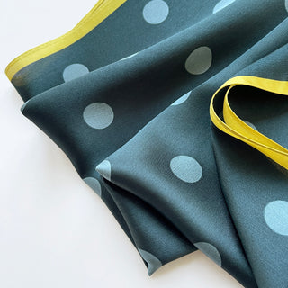 Teal green silk square scarf with light blue polka dots and bright mustard yellow border, elegantly folded showing luxurious sheen and texture