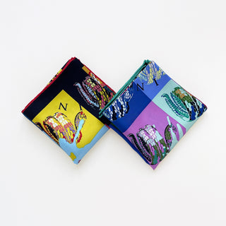 Two folded silk bandanas with colorful snake print designs featuring bold color blocks in blue, red, green, yellow, and purple with hand-rolled edges