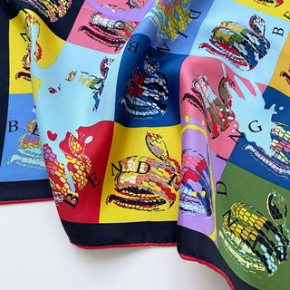 Luxury silk scarf featuring colorful blocks of blue, red, yellow, and pink with artistic snake print designs and letters, bordered in black and red