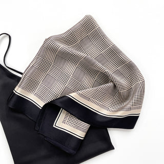 Silk scarf featuring black houndstooth pattern on beige background with solid black border, elegantly folded to display luxurious texture and classic design