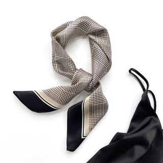 Elegant silk scarf with black houndstooth pattern on beige background, tied in a stylish knot, featuring black border trim and silky texture