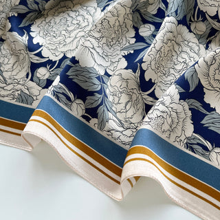 Close-up of luxurious silk scarf with navy blue background and elegant white peony pattern, featuring blue and gold striped border detail