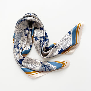 Square silk scarf with elegant peony pattern in blue and white, featuring gold border accents and a lustrous sheen, draped in soft folds