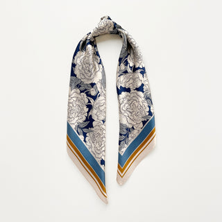 Silk square scarf featuring white peony blooms on navy blue background with blue and gold border details, elegantly draped to showcase pattern