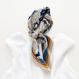 Elegant silk scarf with blue peony pattern and beige floral designs, featuring gold border accents, tied stylishly on white blouse collar