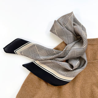 Elegant silk scarf with black houndstooth pattern on beige background, featuring black border trim, draped stylishly over tan fabric