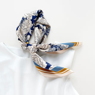 Square silk scarf tied in knot featuring blue background with white peony flower pattern and gold border accents on white surface