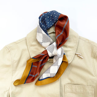 Silk neckerchief with navy geometric, rust, white dotted, and gold patterns tied elegantly around a beige jacket collar with pearl pin