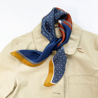 Navy silk neckerchief with geometric pattern, rust accents, and gold border elegantly tied around collar of beige jacket, showcasing luxurious sheen