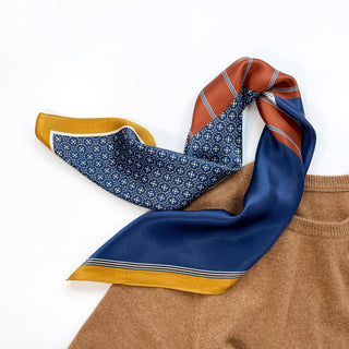 Navy silk neckerchief with geometric patchwork design featuring blue, rust, and gold color blocks, elegantly draped on a tan sweater.