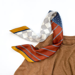 Navy and rust silk neckerchief with geometric patchwork pattern featuring white checkered design, blue medallions, and rust stripes, draped on camel sweater