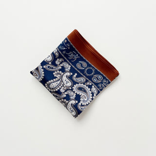 Square navy blue silk scarf with white paisley pattern and rustic brown border, folded neatly on white background