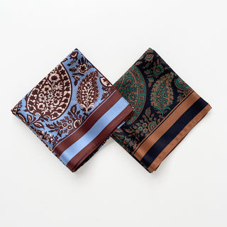 Two folded silk paisley neckerchiefs - one in blue with brown patterns, the other in black with green paisley designs, both featuring elegant borders.