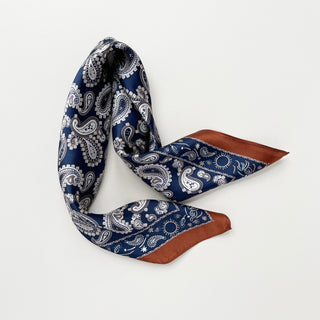 Navy blue silk scarf with white paisley pattern, featuring rustic brown border trim, arranged in casual fold on white background