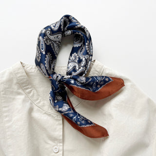 Navy blue silk bandana with white paisley pattern and rustic brown border, styled as a neckerchief on a cream button-up shirt