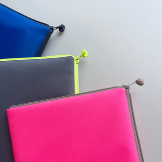 Three recycled RPET iPad mini cases in vibrant colors: bright pink, gray with neon yellow piping, and royal blue, featuring traditional handmade button zipper pulls.