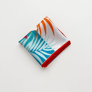 Folded silk bandana with vibrant tropical leaf pattern featuring turquoise and orange palm fronds on white background with red border trim