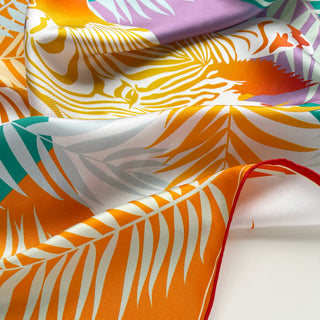 Folded silk scarf with vibrant jungle pattern featuring orange zebra print, purple flowers, and teal leaves on white background with hand-rolled red edge