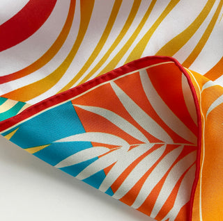 Close-up of double-sided silk scarf featuring vibrant tropical pattern with orange, red, yellow, and turquoise stripes and leaf motifs with hand-rolled edge