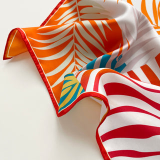 Luxury silk scarf with vibrant tropical print featuring orange, red, and teal palm leaf patterns against white background with hand-rolled edges