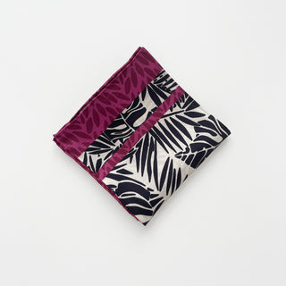Folded silk scarf featuring black tropical foliage pattern on ivory background with magenta leaf-patterned border, displaying hand-rolled edges