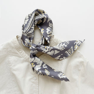 Ash grey silk scarf with white botanical print and yellow accents, styled in a loose knot around the collar of a cream blouse