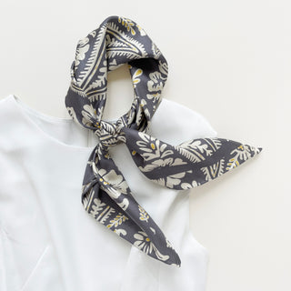Ash grey silk scarf with cream botanical print and yellow accents, styled in a casual neck knot against a white blouse