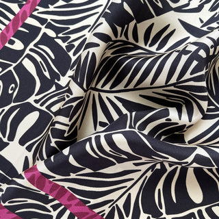 Luxurious silk scarf with black tropical foliage pattern on ivory background, accented by a vibrant magenta border, showing elegant drape and texture