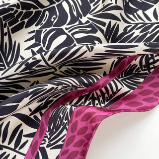 Luxurious silk scarf with black tropical leaf pattern on ivory background, featuring vibrant magenta border with leaf motif, showing silky texture and drape.