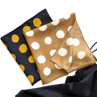 Two artisan silk scarves with polka dot patterns - one navy with gold dots and one ochre with white dots, displayed with hand-rolled edges