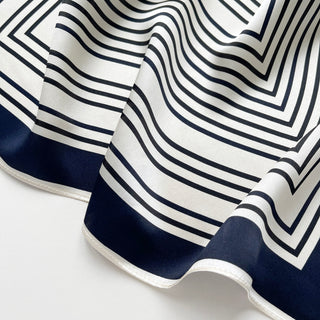 Silk scarf featuring geometric pattern with navy and white stripes, navy border, and shimmering fabric showing elegant drape and texture
