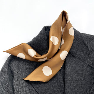 Ochre silk bandana scarf with cream polka dots elegantly draped on a dark gray coat, showcasing its luxurious texture and hand-rolled edges.