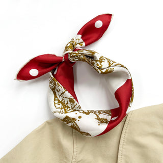 Silk bandana scarf tied in a bow, featuring red polka dot panels and gold paw print design on white background, displayed on beige fabric.