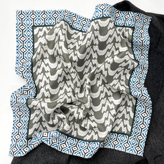 Teal green silk scarf with diamond geometric border pattern surrounding a center of white zigzag designs on gray, displayed on dark fabric