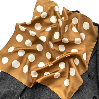 Luxurious ochre silk bandana with cream polka dots, elegantly draped on dark gray fabric showing the scarf's sheen and hand-rolled edges