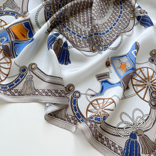 Luxurious silk bandana scarf with equestrian-inspired baroque pattern featuring blue, gold, and brown carriage wheels, chains, tassels, and ornate details on cream background