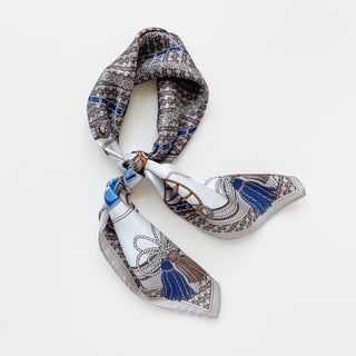 Square silk scarf tied in knot featuring elegant baroque pattern with gray lattice border, blue tassels, equestrian motifs and white center on light background