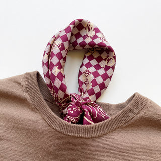 Burgundy baroque check silk scarf with cream patterns tied loosely around the collar of a neutral beige sweater, showing luxurious hand-rolled edges