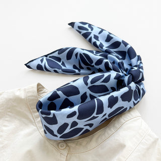 Luxurious silk scarf with navy blue abstract pattern on light blue background, elegantly folded and draped over a cream shirt collar.
