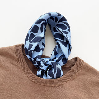Blue Melody silk scarf with navy abstract pattern on light blue background, styled as neck accessory with men's camel sweater, showing luxury texture